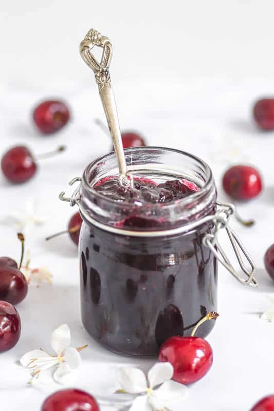 Homemade Spiced Cherry Jam shown with a spoon in the jam jar, next to cherries and flowers 