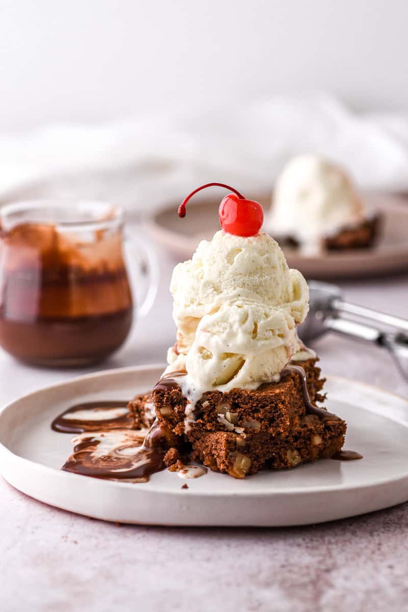 Brownies and ice cream shown stacked on a white plate, the dessert has a cherry place on top of it. 