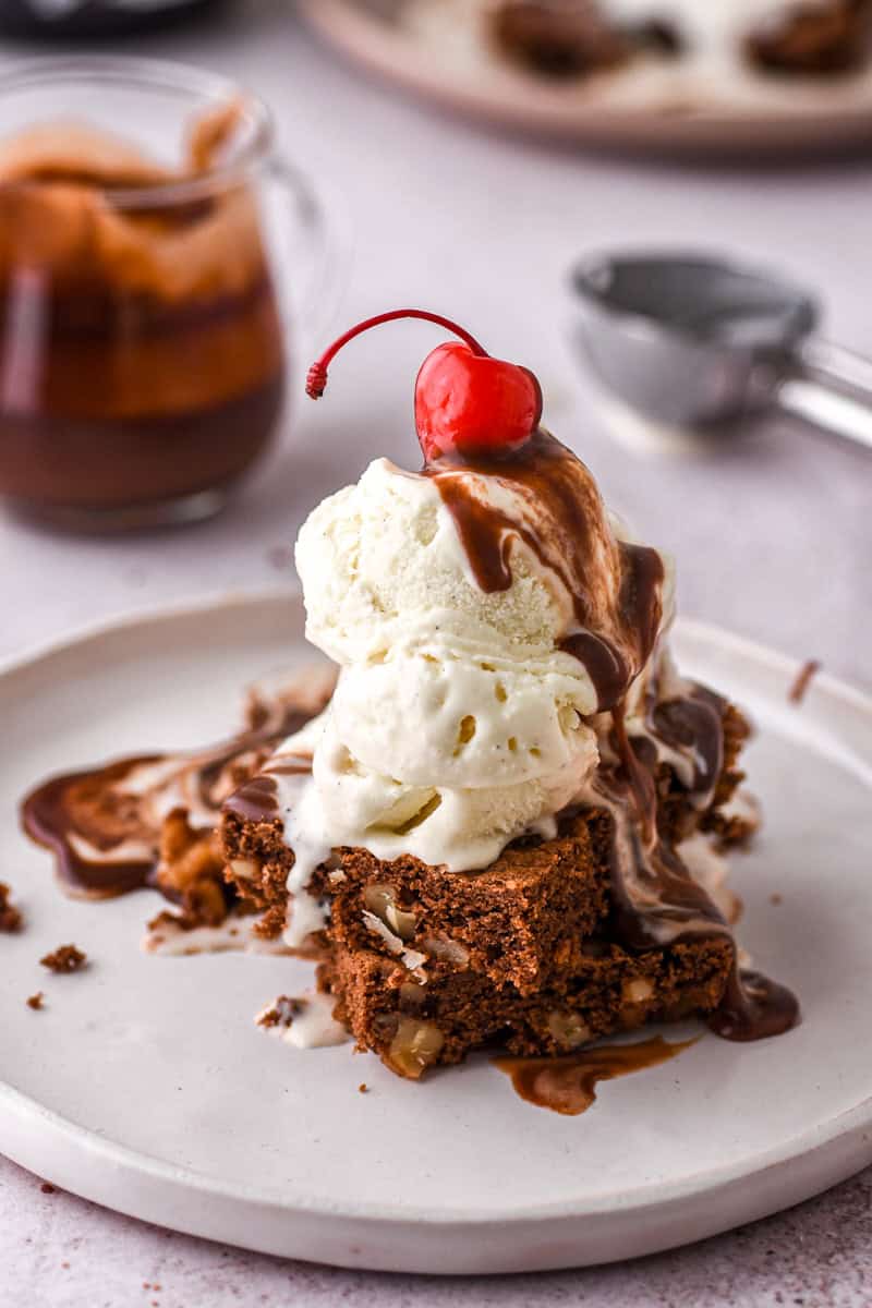 Hot Fudge Brownie Sundae with a cherry on the top shown on a white plate.