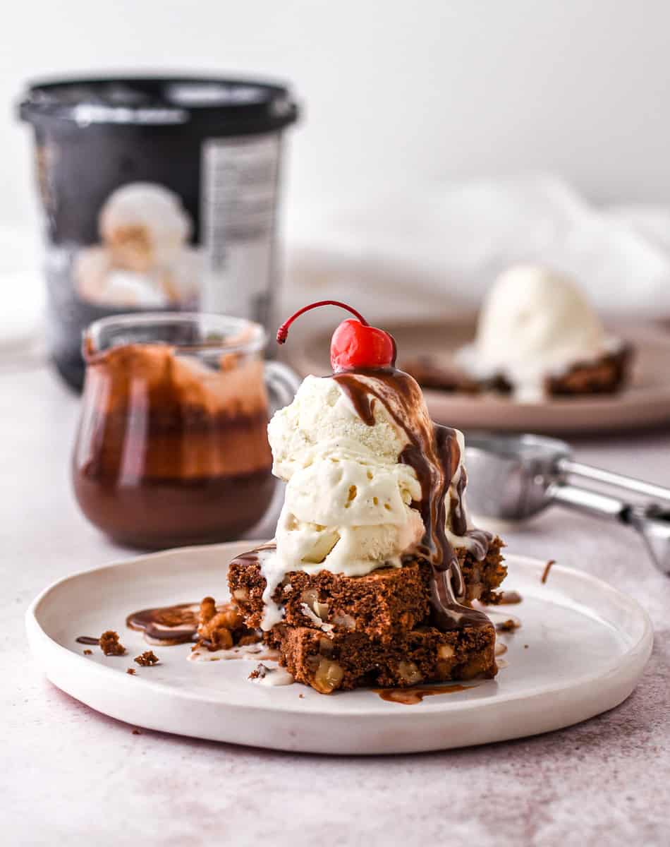 Hot Fudge Brownie Sundae shown on a plate there is a jug of chocolate sauce and a tub of ice cream in the background. 