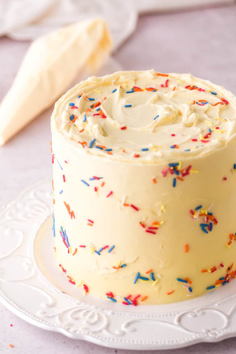 Image shows the decorated cake on a cake plate with a frosting piping bag in the background. 