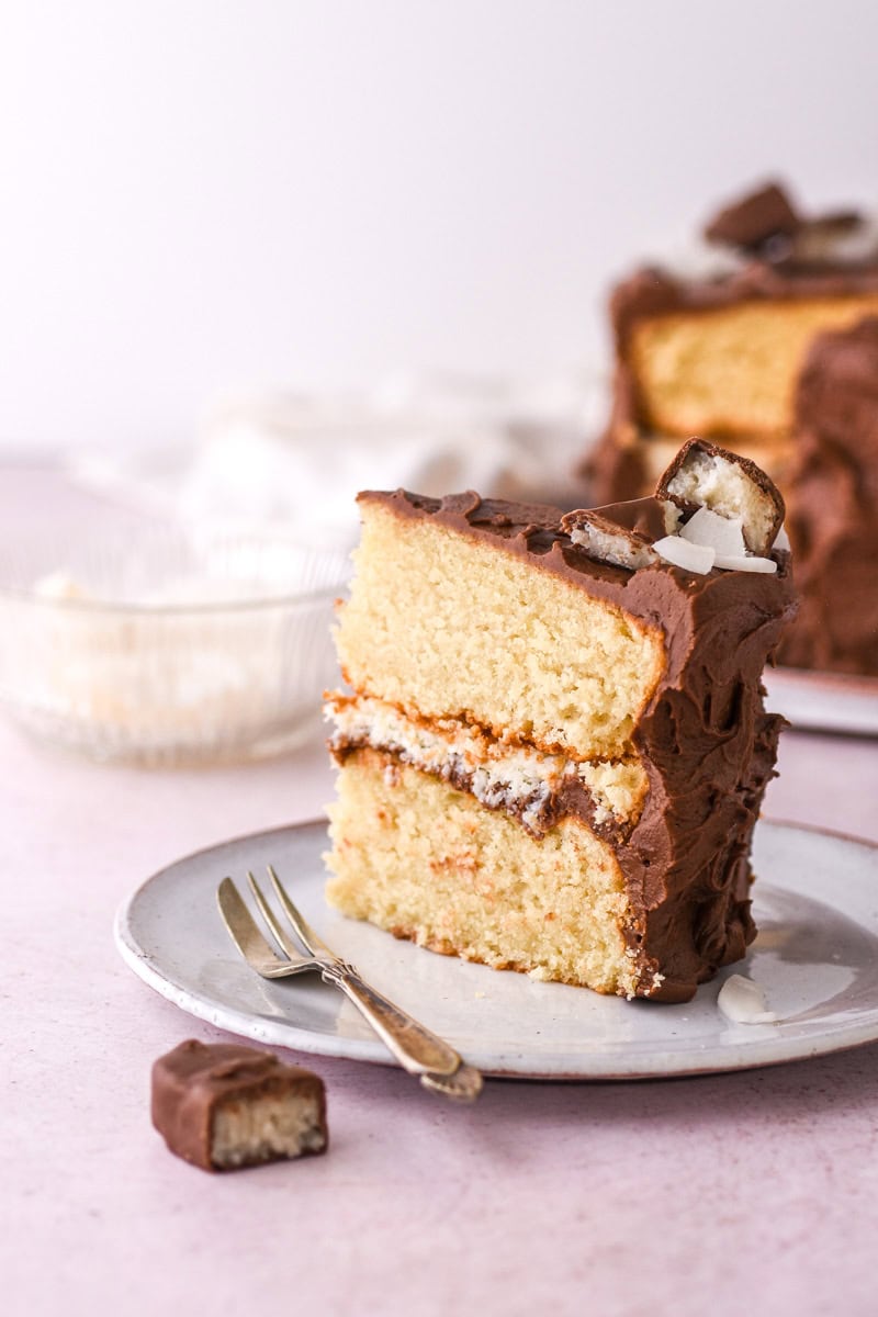 A slice of bounty cake standing upright on a cake plate, there are pieces of coconut and sliced bounty bars on top of the slice. 