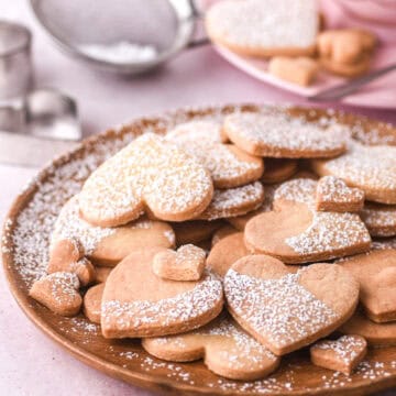 Heart shaped cookies shown on a wooden plate, there is a small sieve and cup of tea in the background.