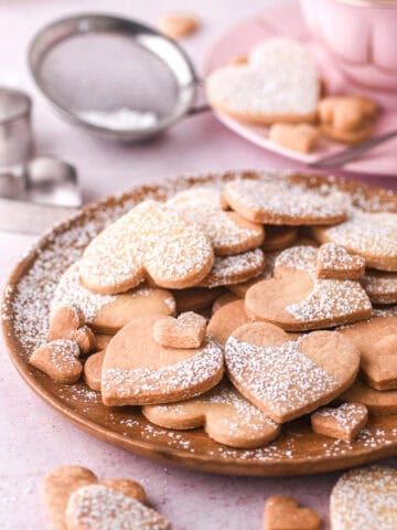 Heart shaped cookies shown on a wooden plate, there is a small sieve and cup of tea in the background.